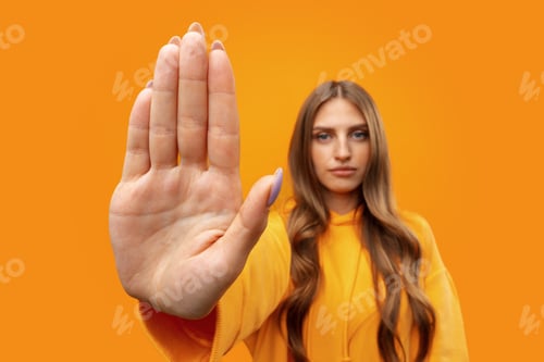 Preview: Young woman standing with outstretched arm showing stop sign against yellow background
