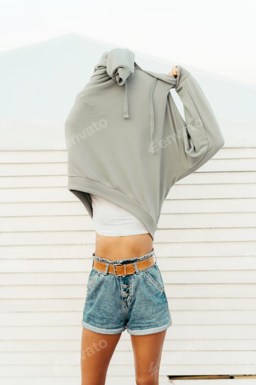Preview: Woman takes off her hoodie while standing on the beach. Daylight. Delicate pastel monochrome