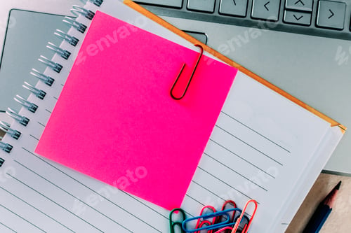 Preview: Top view closeup of a pink sticky note inside of a notebook, secured with a paper clip and a laptop