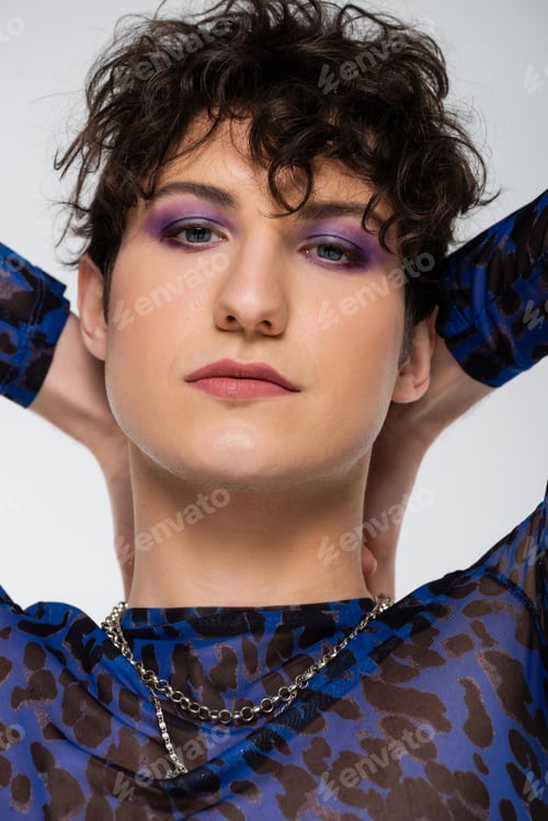 Preview: portrait of brunette queer person with makeup and hands behind head isolated on grey