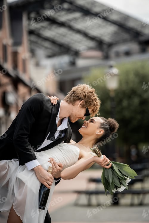 Preview: stylish redhead groom embracing african american bride wit bouquet on city street, outdoor wedding