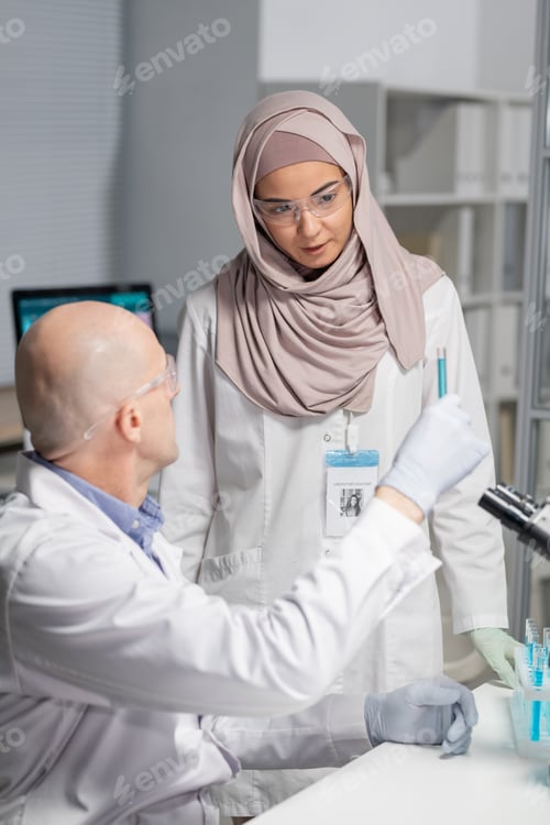 Preview: Young Muslim female virologist in hijab and whitecoat looking at blue liquid in flask held by