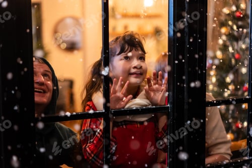 Preview: Adorable child look through the window and admiring first snow flakes falling outside with family.