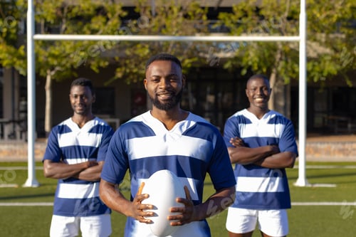Preview: Three African American young male athletes holding a rugby ball, standing on field outdoors