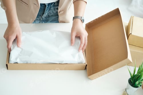 Preview: Asian female clothes shop owner folding a t-shirt and packing in a cardboard parcel box.