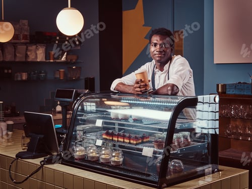 Preview: African barista relaxing after workday with coffee while leaning on the counter at coffee shop.