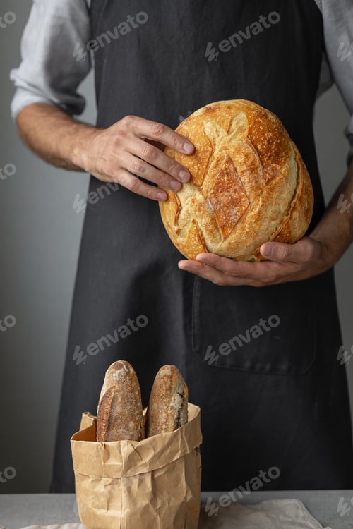 Preview: European male baker in dark apron holding fresh and delicious wheat bread in a paper bag
