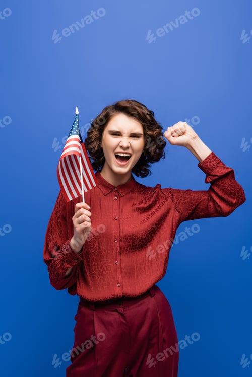 Preview: excited student showing success gesture while holding usa flag and looking at camera isolated on