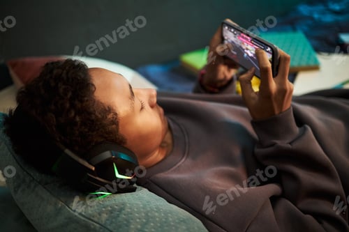 Preview: Teenage Black Boy Wearing Headphones Playing Mobile Game While Relaxing