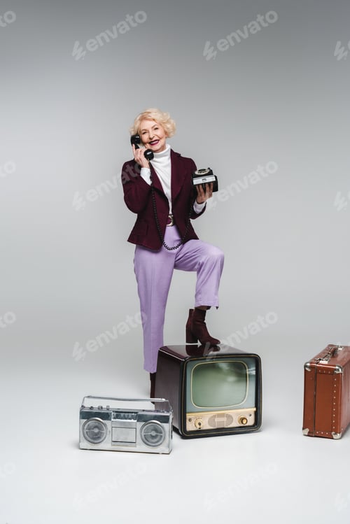 Preview: beautiful senior woman talking by phone while standing on vintage tv on grey