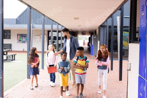 Preview: Diverse, happy male teacher and elementary schoolchildren walking in school corridor, copy space