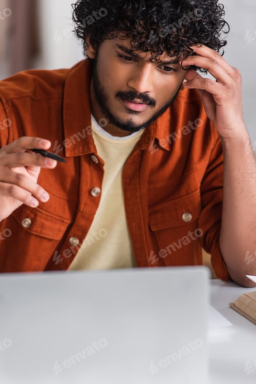 Preview: Focused indian copywriter holding pen near blurred laptop at home