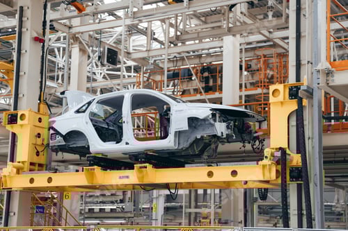 Preview: White car body is suspended on a yellow automated lift within a vast, automotive assembly plant