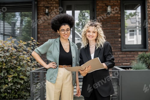 Aperçu: happy realtor with folder and african american woman with hand on hip looking at camera near house