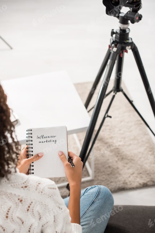 Preview: cropped view of curly african american girl holding notebook with influencer marketing lettering at