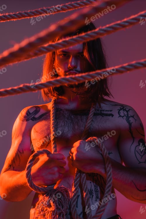 Preview: Young tattooed model looking at camera near blurred ropes on purple background with light