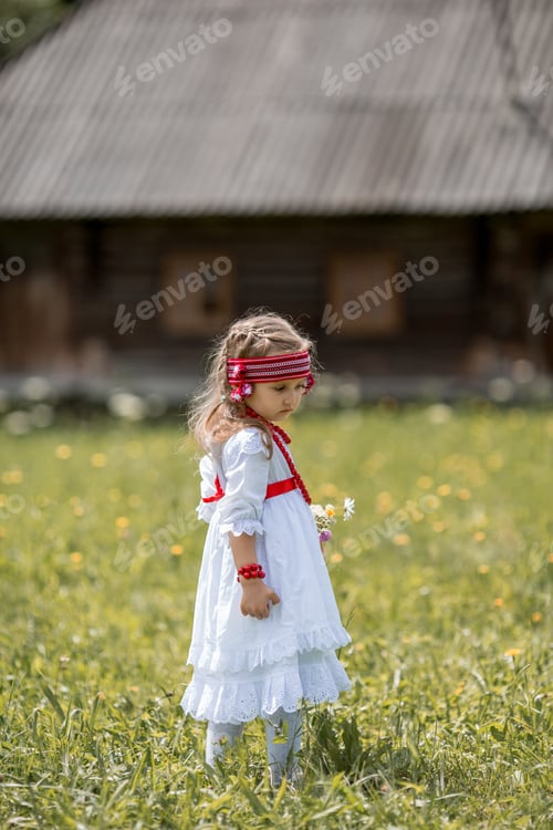 Preview: A little girl in a white dress and national Ukrainian accessories near a village house
