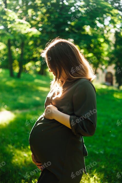 Preview: Portrait of young pregnant woman outdoors looking at her belly in a park