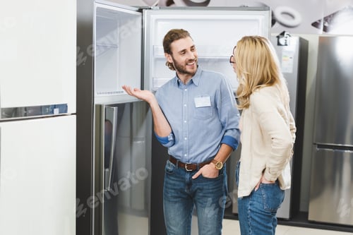 Preview: smiling consultant pointing with hand at fridge and talking with woman in home appliance store