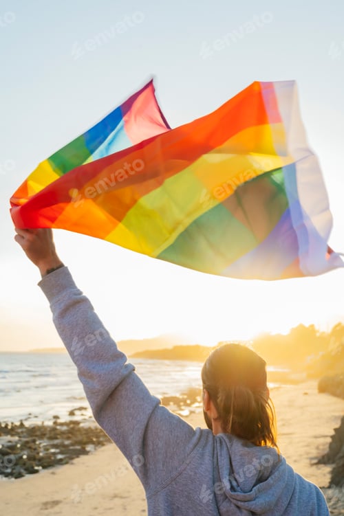 Preview: Man raising LGBT rainbow flag to the sky on the beach