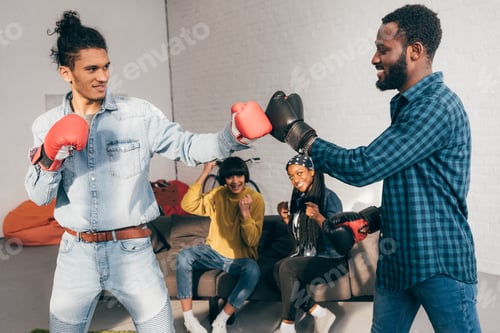 Preview: multicultural male friends doing sparring in boxing gloves and two women cheering up them