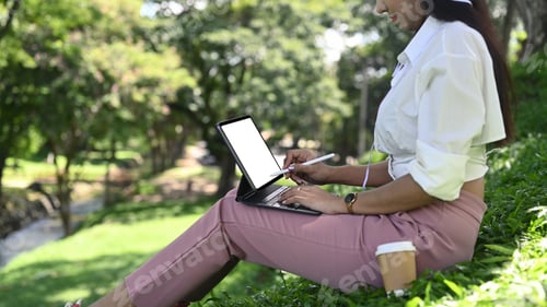 Preview: Young woman sitting in public park and working with computer tablet.