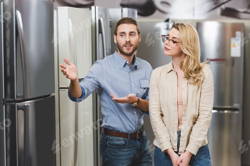 Preview: consultant pointing with hands at fridge and talking with woman in home appliance store