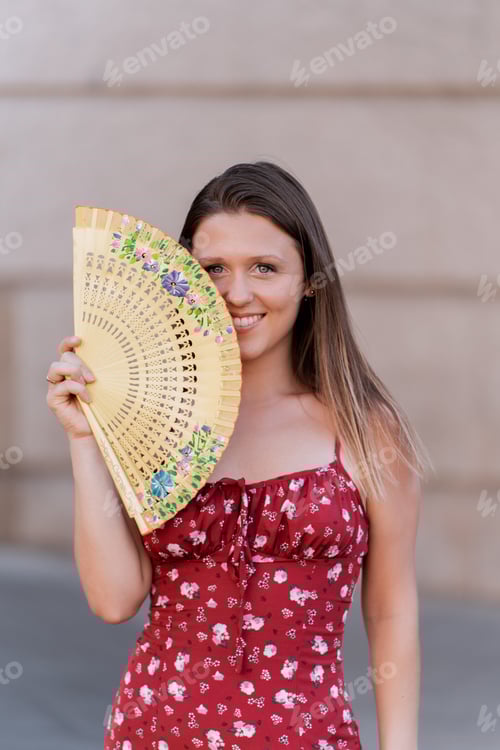 Preview: Woman looking at camera and smiling while holding a hand fan outdoors.