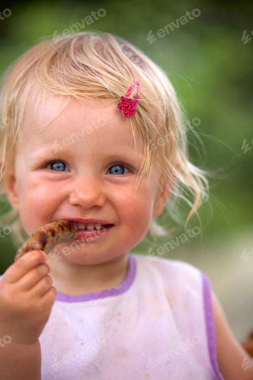 Preview: Portrait of blond little girl with bib and pink haipin eating sausage
