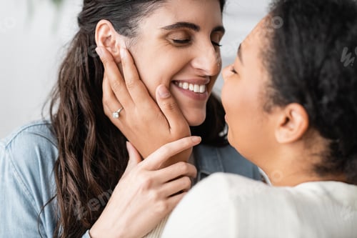 Preview: overjoyed multiracial woman with engagement ring on finger hugging smiling girlfriend