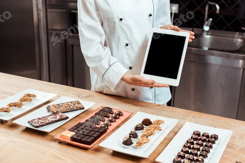 Preview: cropped view of chocolatier in holding digital tablet with blank screen near tasty chocolate candies