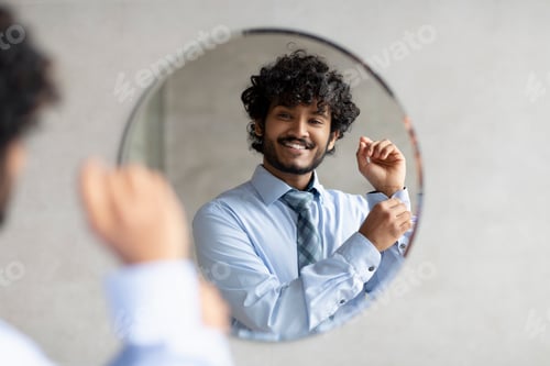 Preview: Happy bearded indian businessman standing in bathroom in front of mirror and buttoning up shirt