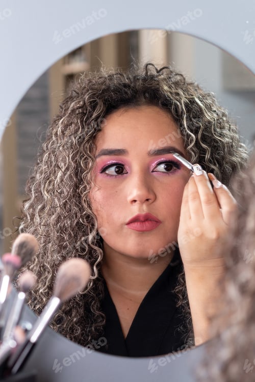 Preview: Vertical photo of latin woman with curly hair applying eyebrow makeup in front of a mirror
