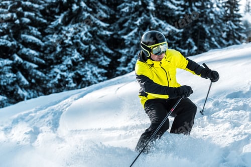Preview: skier in goggles and helmet holding ski sticks and skiing near firs
