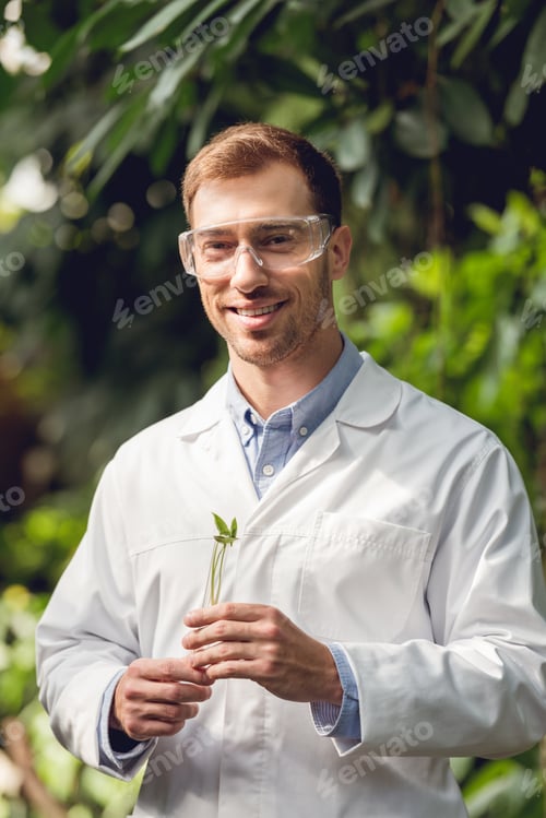 Preview: happy scientist in white coat and goggles holding flask with plant sample in orangery