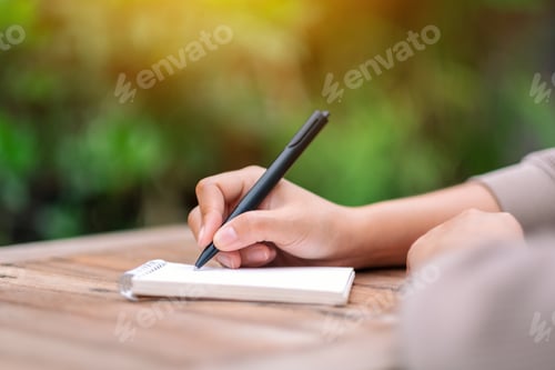 Preview: Closeup image of a woman writing on blank notebook on wooden table in the outdoors