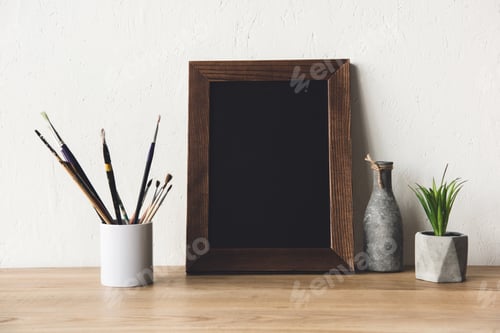 Preview: close up view of empty photo frame, vase and paintbrushes on wooden tabletop