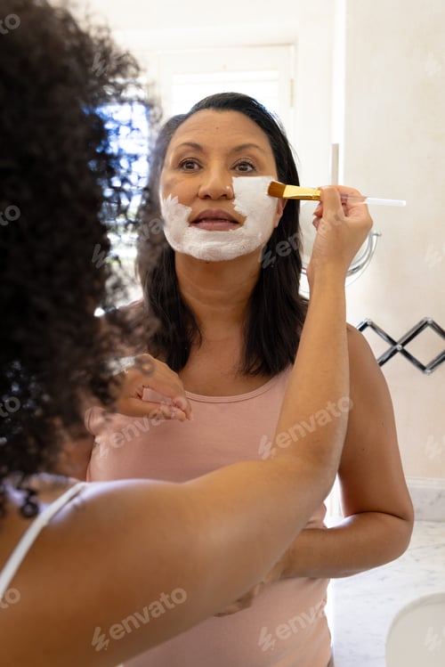 Preview: Mature biracial woman and daughter use facial masks, looking in mirror