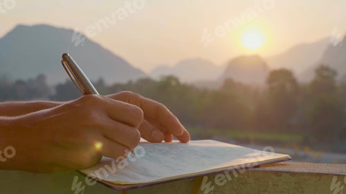 Preview: Closeup female hands writing travel notes in diary during sunrise with beautiful sun light and