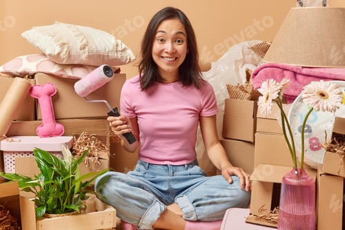 Preview: Happy Asian woman holds paint roller to improve apartment relocates to new flat surrounded by cardbo