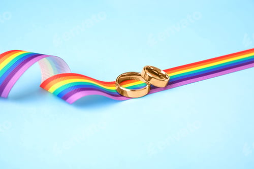 Preview: Wedding rings and rainbow ribbon on color background. Gay symbol