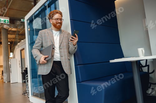 Preview: Smiling businessman in eyeglasses with laptop standing on modern office background and use phone