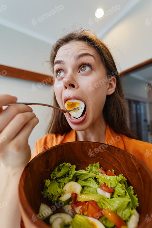 Preview: Young woman excitedly eating a fresh salad with a wooden bowl in a bright kitchen, showcasing