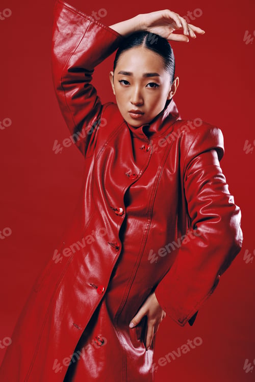 Preview: Fashionable asian brunette woman in red leather jacket posing with hands on hips against red