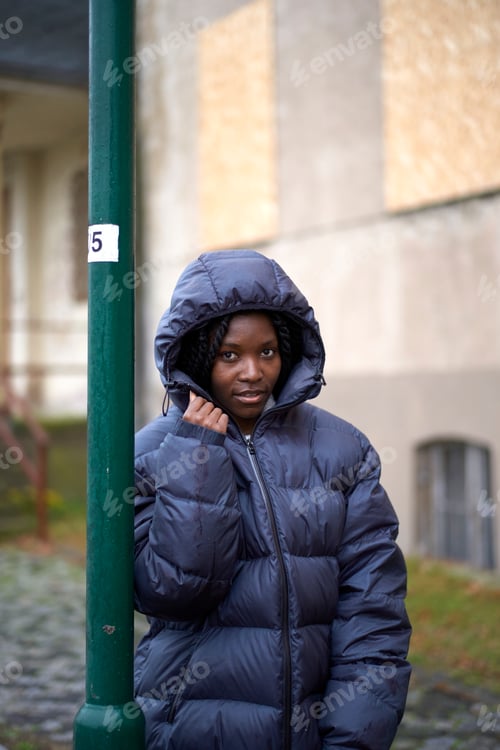 Preview: Young woman in a puffy jacket leaning against a green pole on a cloudy day, Brandenburg, Germany