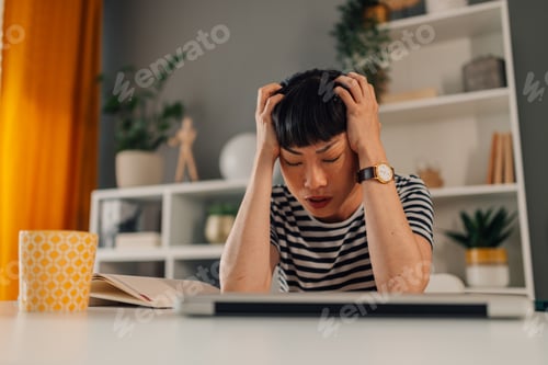 Preview: Asian woman frustrated at work, sitting with hands on her head, stressed.