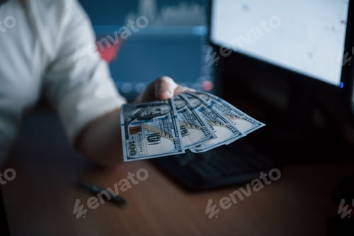 Preview: Bearded man in white shirt holds money in the office with multiple computer screens in index charts
