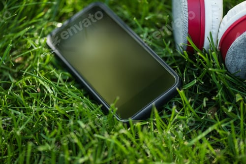 Preview: close up view of smartphone with blank screen and headphones on green grass