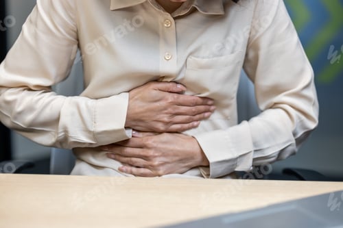 Preview: Woman in beige blouse experiencing stomach pain while seated at office desk during work hours