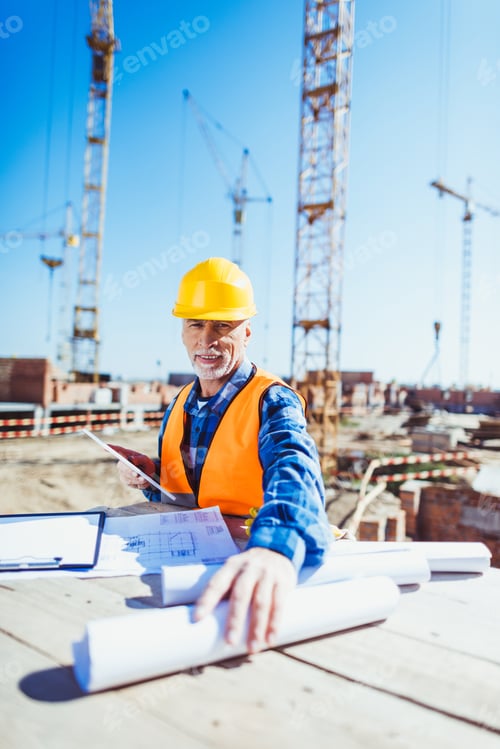 Preview: Construction worker in reflective vest and hardhat, holding rolls with building plans and looking at